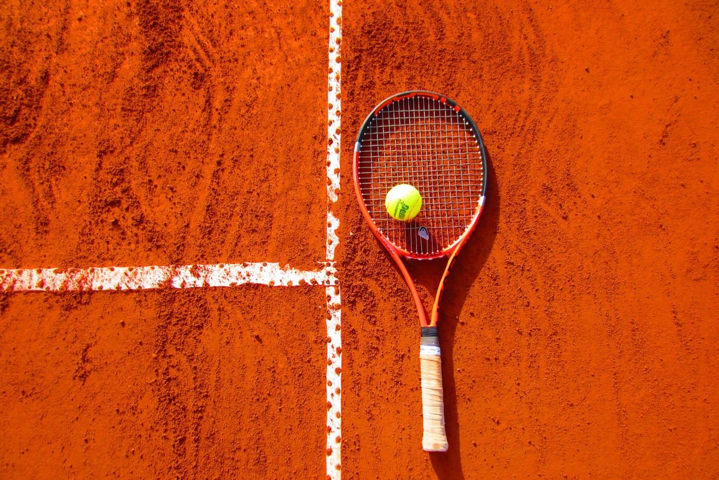 A tennis racket and ball resting on a red clay court beside the white baseline